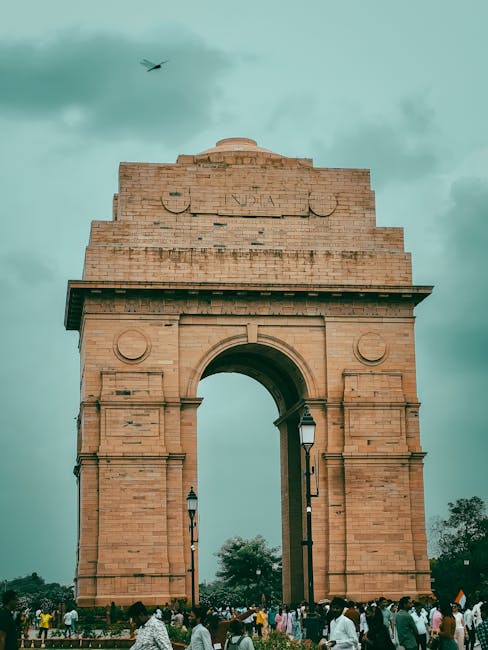 A view of the iconic India Gate in New Delhi with a crowd gathered underneath and a cloudy sky.